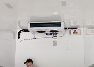 Man in a cap using a drill on a white wall inside a utility trailer or workshop, with metal diamond-plate trim and equipment mounted above him