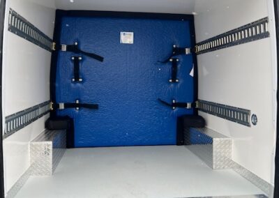 Interior of white cargo van with blue rear bulkhead and metal tie-down rails along the walls and floor edges for securing cargo.