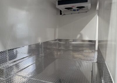 Interior of a stainless steel trailer with a diamond-plate floor and a ceiling air conditioning unit mounted on the wall/ceiling area.