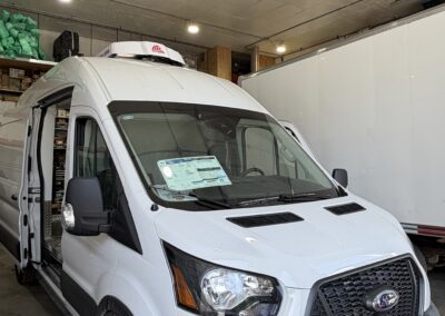 White cargo van inside a garage with the driver's side door open and a roof cargo box visible on top; shelves and storage along the wall in the background.
